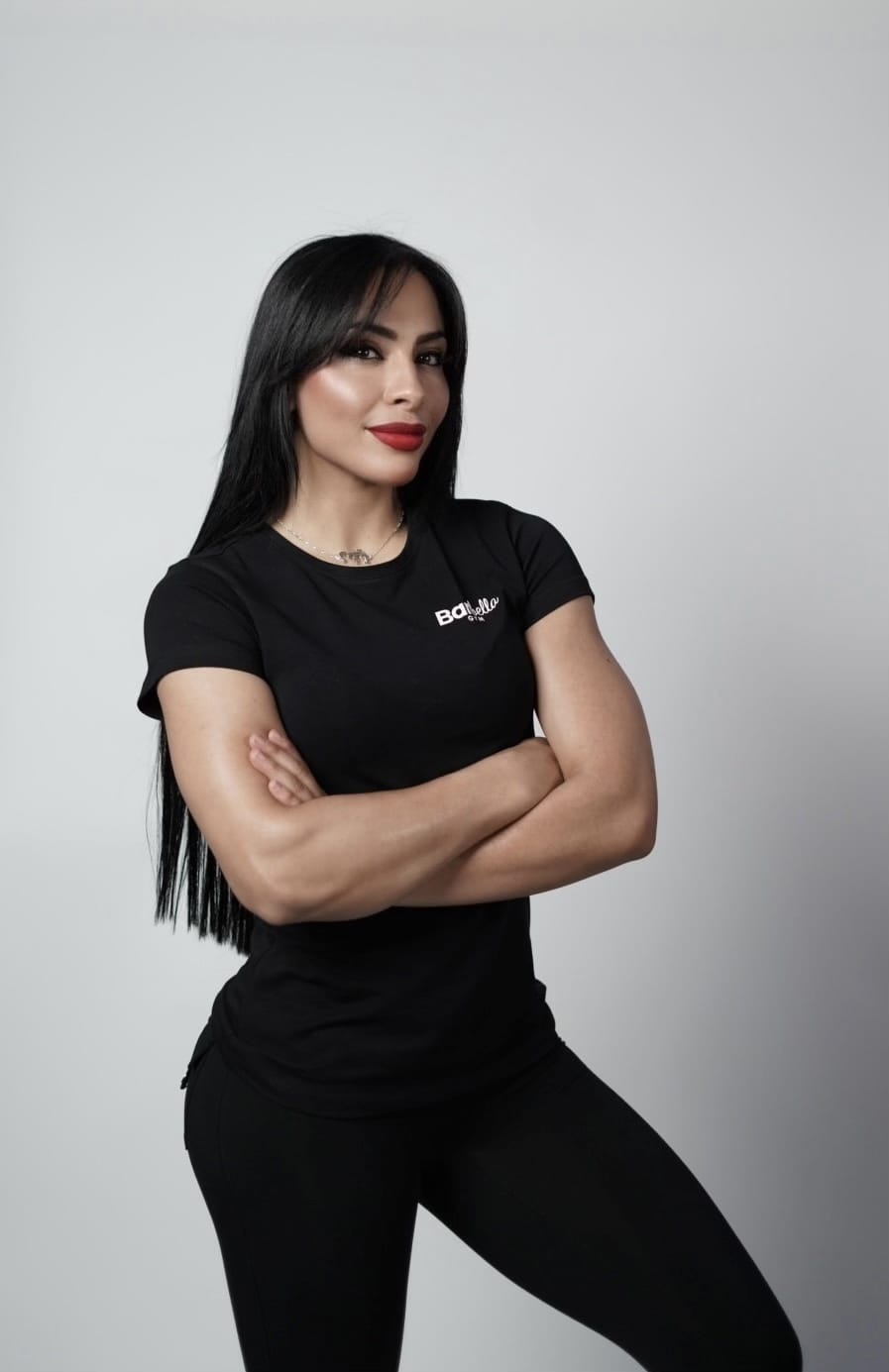 A woman with long black hair, red lipstick, and a black fitted t-shirt with "Bot Life" text stands against a plain light gray background, arms crossed, smiling confidently. She wears black pants and has a tattoo visible on her necklace.