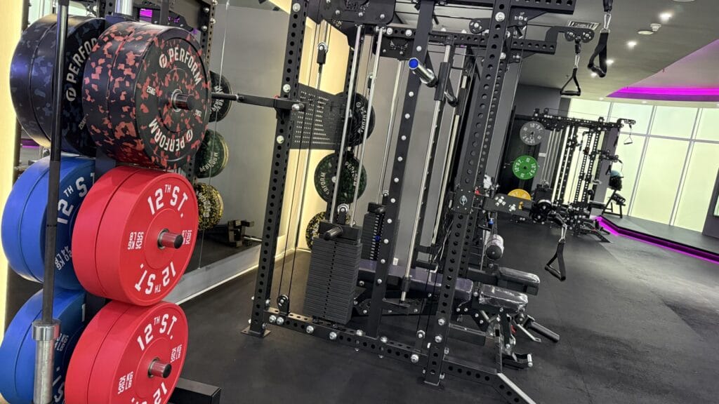 A modern gym features a rack of colorful weight plates (red, blue, black) beside a cable machine. Other gym equipment, benches, and weights line the spacious, brightly lit room with black floors and purple accent lighting.