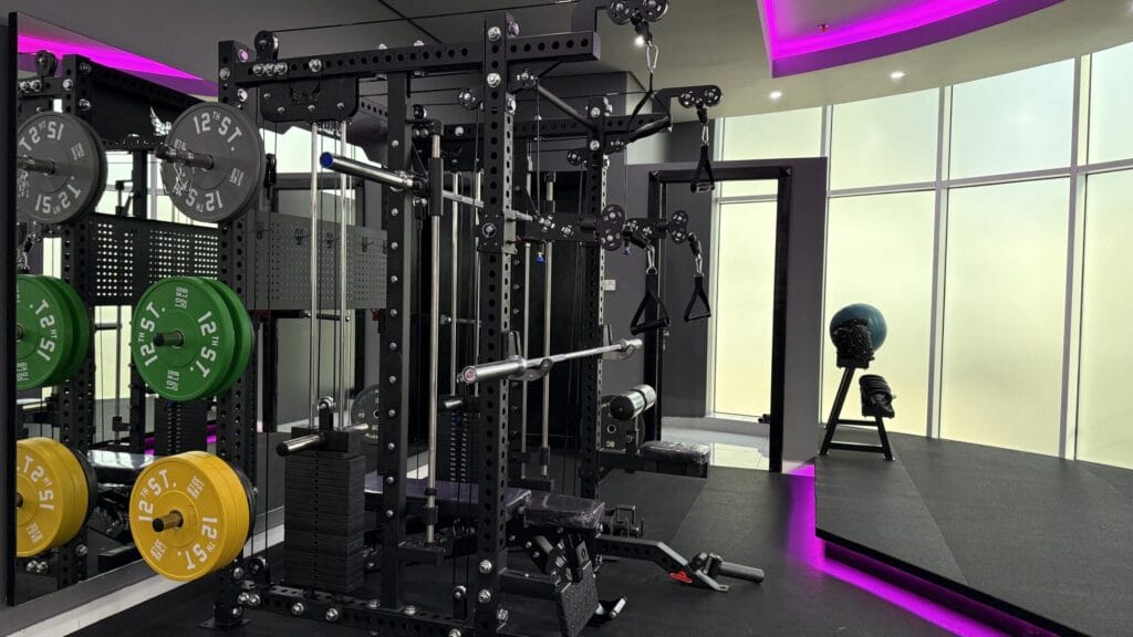 A modern gym features weightlifting equipment with green, yellow, and gray weight plates. The room has black machines, neon purple lighting, large frosted windows, and a human head sculpture on a stand near the back wall. The floor is sleek and dark.