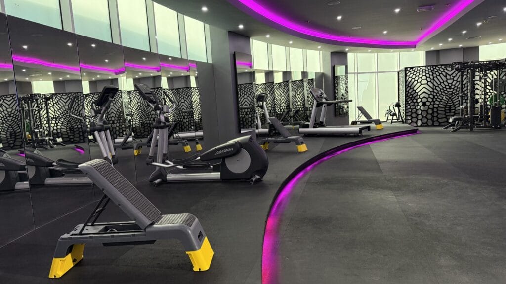 A modern gym with exercise equipment including treadmills and benches, mirrored walls, black patterned walls, and a curved strip of purple LED lighting along the ceiling and floor, creating a vibrant, sleek atmosphere. The space is clean and well-lit.