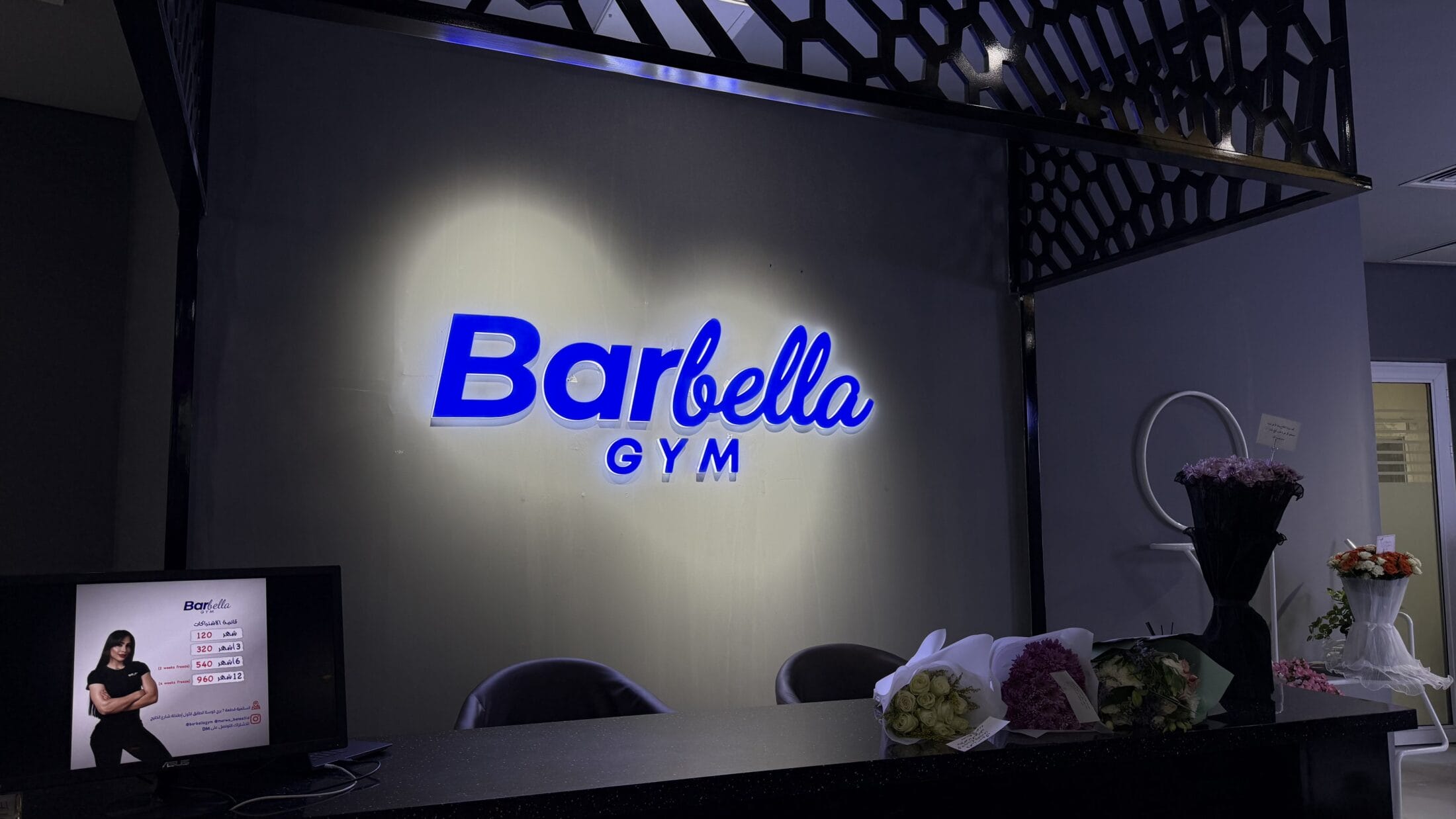 The reception area of Barbella Gym features a glowing blue logo on a gray wall, bouquets of flowers on the counter, a small display screen with a woman in gym attire, and a dark geometric ceiling fixture above. The space has a modern and welcoming design.
