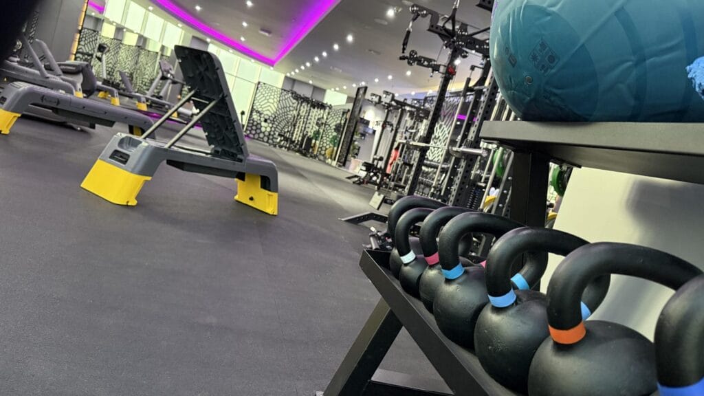A gym with a row of black kettlebells on a rack in the foreground, a blue exercise ball above them, benches with yellow bases, strength machines, and racks in the background. The ceiling features purple LED lighting.