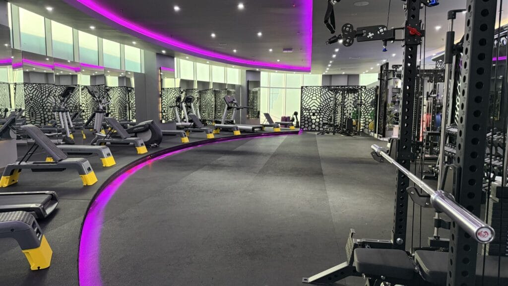 Modern gym with treadmills, weight machines, and benches, all arranged neatly. The curved ceiling and floor feature glowing purple LED lights. Walls have geometric black-and-white patterns, and large mirrors and windows make the space bright and spacious.