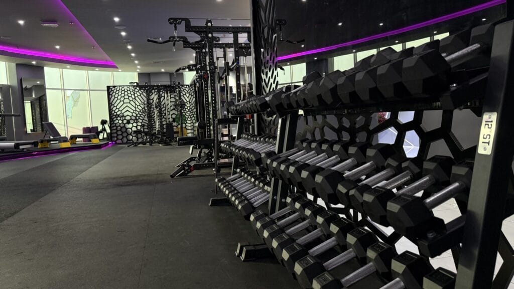 A modern gym with rows of hexagonal dumbbells on racks, exercise machines, and weight benches. The space features geometric wall designs, large windows, and purple LED accent lighting, creating a sleek and contemporary atmosphere.